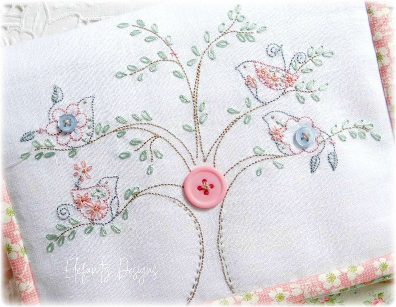 May include: Embroidered white fabric with a tree design featuring three birds, pink and blue flowers, and a pink button. The text "Elfauts Designs" is embroidered at the bottom.