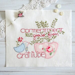 May include: Embroidered white fabric with the text "Contentment grows from gratitude" in red thread. A pink fabric heart with a rose design is stitched below the text. A blue bird with white polka dots is stitched to the left of the heart.