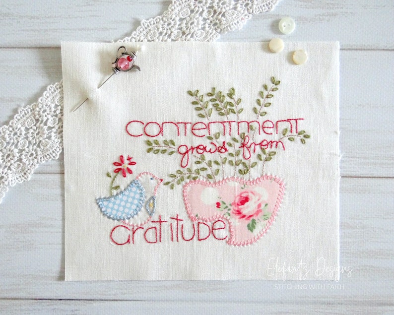 May include: Embroidered white fabric with the text "Contentment grows from gratitude" in red thread. A pink fabric heart with a rose design is stitched below the text. A blue bird with white polka dots is stitched to the left of the heart.