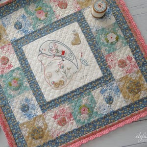 May include: A small, square quilt with a hand-stitched design of a bird and a pear in the center. The quilt is made of various floral fabrics in shades of blue, pink, and green, and has a crocheted border.