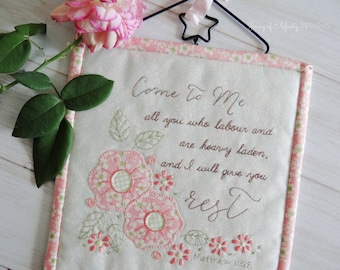 Puede incluir: Una pared colgante de tela blanca con bordado floral rosa y el texto "Venid a mí, todos los que estáis cansados y cargados, y yo os haré descansar Mateo 11:28".