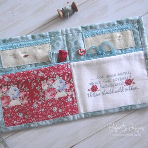 May include: A blue and red fabric sewing roll-up with multiple pockets and compartments. The roll-up has a white fabric panel with the embroidered text "...for good works, which God prepared beforehand that we should walk in them." The roll-up is labeled "Elefants Designs STITCHING WITH FAITH".