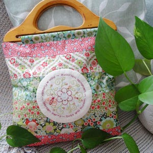 May include: A patchwork fabric tote bag with a wooden handle. The bag features a circular embroidered patch with the text "Welcome to our garden party - pick a flower and bloom".