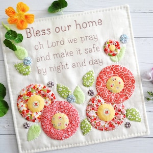 Könnte beinhalten: Ein weißer Wandbehang aus Stoff mit floralem Muster und dem Text "Bless our home oh Lord we pray and make it safe by night and day".