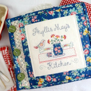 May include: A blue floral fabric covered cookbook with a white embroidered cover that says "Phyllis May's Kitchen" with a bird and flowers.