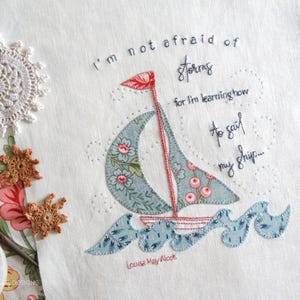 May include: A white fabric with blue and red embroidery of a sailboat with the text "I'm not afraid of storms for I'm learning how to sail my ship... Louisa May Alcott"