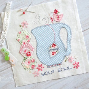 May include: A blue and white teapot with a tea bag that says "revive" and a small bird perched on top. The teapot is surrounded by pink and green floral fabric scraps and the text "revive your soul" is stitched below.