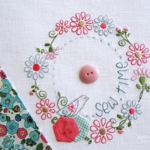 Può includere: Design floreale ricamato con un bottone rosa. Il design presenta una ghirlanda di fiori rosa, rossi e blu, con le parole "Sew Time" ricamate in blu. Un piccolo uccello è incluso nel design.