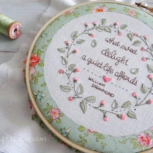 Puede incluir: Arte de bordado en un aro con una corona floral y la cita "Qu&eacute; dulce deleite ofrece una vida tranquila" de William Drummond. El aro est&aacute; rodeado de una tela floral.