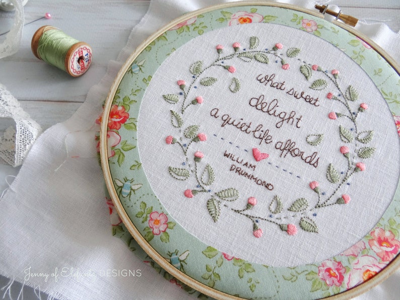 Puede incluir: Arte de bordado en un aro con una corona floral y la cita "Qu&eacute; dulce deleite ofrece una vida tranquila" de William Drummond. El aro est&aacute; rodeado de una tela floral.