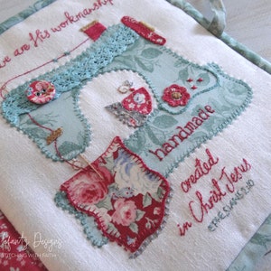May include: A white fabric book with a blue and green sewing machine design. The book has red and white floral fabric accents and the text "For we are His workmanship" and "handmade created in Christ Jesus Ephesians 2:10".