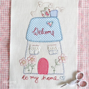 May include: Embroidered white fabric with a blue and white house design. The house has a red door and two windows. The words "Welcome to my home" are embroidered below the house. There are two birds on the roof of the house.