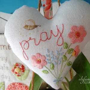 以下が含まれることがあります： ピンクの糸で「祈る」と刺繍された、白いハート型の布製のクッション。花も刺繍されています。