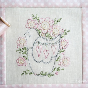 May include: Embroidered design of a bird holding a bouquet of pink flowers with a string of hearts hanging from its beak. The design is stitched on a white linen fabric.