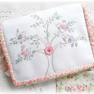 May include: A white fabric pouch with a pink and white floral trim. The pouch is embroidered with a tree with three birds and pink and blue buttons.