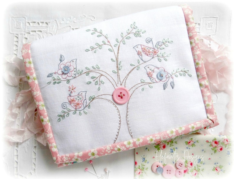 May include: A white fabric pouch with a pink and white floral trim. The pouch is embroidered with a tree with three birds and pink and blue buttons.
