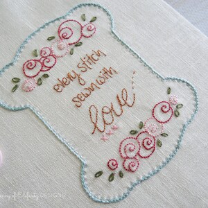 Peut inclure: Tissu brod&eacute; en forme de bobine avec le texte "every stitch sewn with love" en fil brun. La bobine est d&eacute;cor&eacute;e de roses roses et rouges et de feuilles vertes.