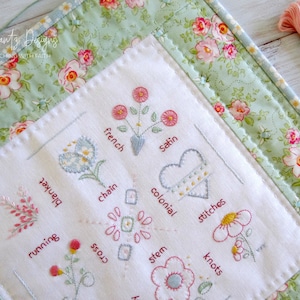 May include: A white fabric sampler with various embroidery stitches, including running stitch, blanket stitch, cross stitch, chain stitch, French knots, satin stitch, colonial stitch, and stem stitch. The fabric is surrounded by a green floral border.