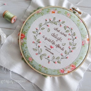 Puede incluir: Un aro de bordado blanco con un borde de tela verde y floral. El bordado presenta una cita en hilo marr&oacute;n: "What sweet delight a quiet life affords" con un coraz&oacute;n y el nombre "William Drummond" debajo.
