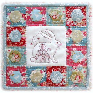 Może przedstawiać: Biały kwadratowy kawałek materiału z ręcznie wyszytym królikiem trzymającym doniczkę. Materiał jest otoczony patchworkiem kolorowych kwadratowych kawałków materiału z kwiatowymi wzorami.