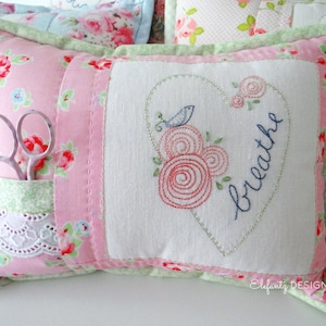 May include: A pink and green cushion with a white fabric panel featuring a heart with a bird and the word "breathe" embroidered in blue thread. The cushion has a pocket on the side with a pair of scissors inside.