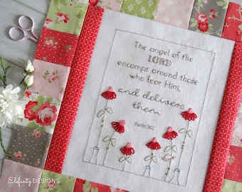 May include: A white fabric square with a brown embroidered quote: "The angel of the LORD encamps around those who fear Him, and delivers them. Psalm 34:7". The fabric is surrounded by a red and white patchwork border.