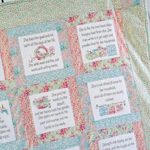 Op de afbeelding: Een patchworkdeken met bloemmotieven in roze, blauw en groene tinten. Elk vierkant heeft een geborduurd citaat over de deugden en het werksethos van een vrouw. De citaten zijn: "Ze doet hem goed en schaadt hem niet alle dagen van haar leven. Ze zoekt wol en vlas en werkt met bereidwillige handen." "Ze is als de koopvaardijschepen, die voedsel van ver brengen. Ze staat op terwijl het nog nacht is en zorgt voor voedsel voor haar huishouden." "Ze legt haar handen op de spinrok, en haar handen houden de spil vast. Ze opent haar hand voor de armen en strekt haar handen uit naar de behoeftigen." "Ze is niet bang voor sneeuw voor haar huishouden, want ze zijn allemaal dubbel gekleed." "Kracht en waardigheid zijn haar kleding, en ze lacht om de"