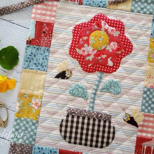 May include: A quilted wall hanging featuring a red flower with a yellow centre, green leaves, and a brown and white checked pot. Two brown and yellow butterflies are on either side of the flower. The background is a white fabric with brown and white stripes.