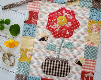 May include: A quilted wall hanging featuring a red flower with a yellow center, green leaves, and a brown and white checkered pot. Two brown and yellow butterflies are on either side of the flower. The background is a white fabric with brown and white stripes.