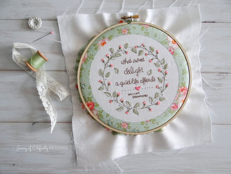 Puede incluir: Un aro de bordado verde y blanco con un dise&ntilde;o floral y la cita "Qu&eacute; dulce deleite ofrece una vida tranquila - William Drummond" bordada en hilo rosa.