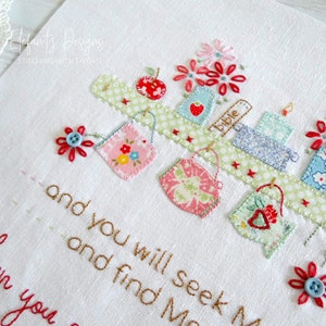 Puede incluir: Tela blanca bordada con un estante que contiene varios art&iacute;culos, incluyendo un libro etiquetado como "bible", una manzana roja y una vela. El estante est&aacute; decorado con tela verde y blanca y estrellas rojas. El texto "and you will seek M and find Me when you" est&aacute; bordado en hilo marr&oacute;n debajo del estante.