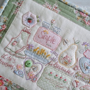 May include: A white fabric square with colorful embroidered designs, including a pear, a heart, a flower pot, and a sewing kit. The text "Simple Pleasures" and "Life's Treasures" are embroidered on the fabric.