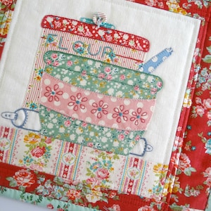 May include: A quilted kitchen pot holder with a floral design. The center features a patchwork design of a bowl, rolling pin, and a container labeled "FLOUR".