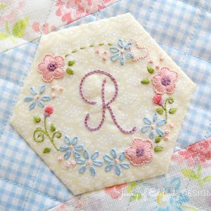 Pode incluir: Um patch bordado hexagonal com um design floral e a letra "R". O patch apresenta flores rosa e azuis, folhas verdes e uma letra bordada roxa sobre um fundo creme. O patch está sobre um tecido acolchoado.
