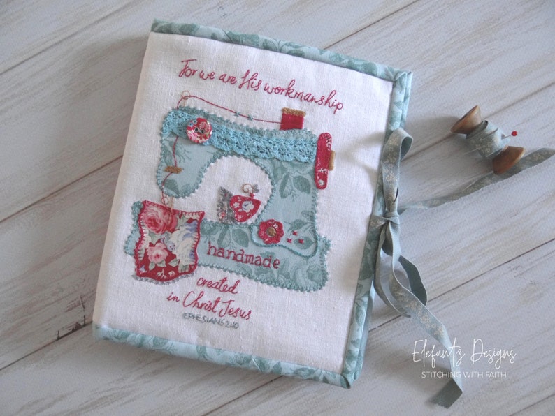 May include: A white fabric book cover with a blue and red embroidered sewing machine design. The cover has a blue ribbon tied around it and the text "For we are His workmanship handmade created in Christ Jesus Ephesians 2:10" is embroidered on the front. The book cover is on a white wooden surface.