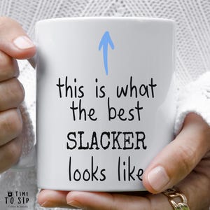 May include: White ceramic mug with black text that reads "This is what the best slacker looks like" with a blue upward arrow above the text.
