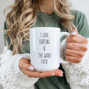 Puede incluir: Taza de cerámica blanca con el texto "I LOVE SURFING & THE WORD FUCK" en negro. La taza tiene un asa grande y la sostiene una persona que lleva anillos y un suéter blanco.