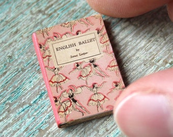 May include: A miniature book titled "English Ballet" with a pink cover featuring illustrations of ballerinas. The book is held between fingers, showcasing its small size. The background is a distressed, light blue wooden surface.