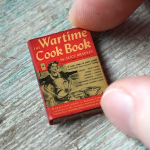 Puede incluir: Un libro de cocina en miniatura titulado "The Wartime Cook Book" de Alice Bradley. La portada es roja con letras doradas y una ilustración de una mujer. El libro se sostiene entre dos dedos sobre una superficie de madera azul claro desgastada.