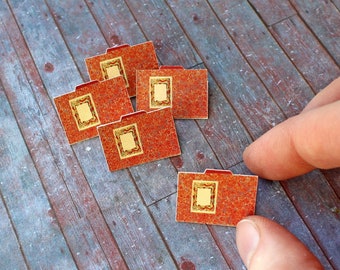 May include: Miniature red folders with gold frames are arranged on a weathered wooden surface. The folders have a textured appearance and a small tab at the top. A hand is reaching towards the folders.