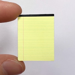 May include: A miniature yellow legal pad with horizontal blue lines and a vertical red margin line. The notepad features a black binding strip at the top edge, designed in a classic office stationery style for small-scale note-taking purposes.