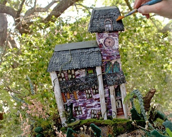 May include: A miniature haunted house with a black roof and purple and white walls. The house is surrounded by green foliage and small trees. A hand is painting the roof of the house with a paintbrush.