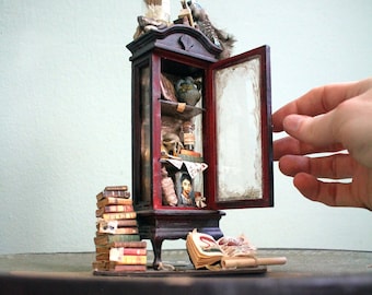 May include: Miniature wooden cabinet with a red interior and glass door, filled with tiny books, bottles, and a portrait. A stack of miniature books and an open book are next to the cabinet. A hand is opening the cabinet door.