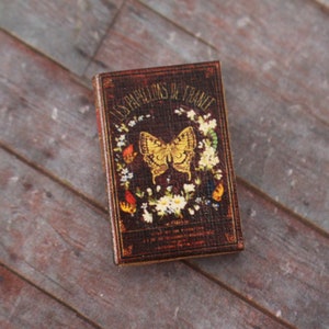 May include: A miniature book with a dark brown cover featuring the title "LES PAPILLONS DE FRANCE" and a gold butterfly illustration surrounded by flowers. The book has a vintage aesthetic and is set against a wooden background.