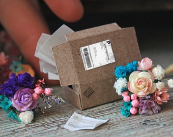 May include: Three miniature flower bouquets with pink, purple, white, and blue flowers. The bouquets are arranged on a wooden surface with a small brown cardboard box and a piece of paper.