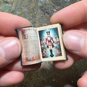 Peut inclure: Un livre miniature avec une couverture rouge et or. Le livre est ouvert sur une page avec une illustration colorée d'un soldat de jouet en uniforme rouge. Le texte sur la page est "Casse-Noisette".