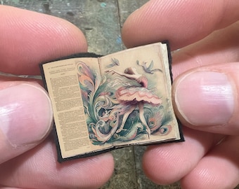 May include: A small, open book with a vintage illustration of a ballerina dancing among swirling floral patterns and birds. The book has a black cover and aged pages.