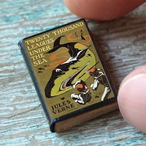 May include: Miniature book titled "Twenty Thousand Leagues Under the Sea" by Jules Verne. The cover features a gold and black illustration of a submarine and divers. The book is held between two fingers, showcasing its small size.