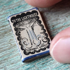 May include: A miniature book titled "TO THE LIGHTHOUSE" by Virginia Woolf. The book has a cream-colored cover with a black and gray illustration of a tree and lighthouse. The book is held between two fingers.