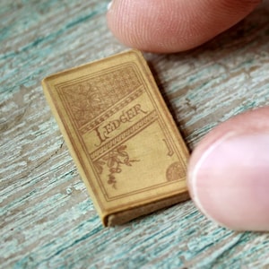 May include: A miniature book labeled "Ledger" with a tan cover and decorative floral elements. The tiny book is held between two fingers, showcasing its small size. The background is a distressed, teal-colored wooden surface.